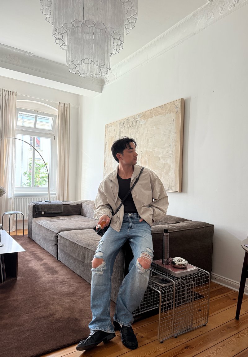 Homme portant un jean déchiré et une veste claire assis sur un canapé gris dans un salon lumineux avec une grande peinture et une table d'appoint en fil métallique.