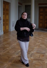 Veste noire zippée à col montant, portée par-dessus un haut transparent orné de dentelle, associée à un pantalon beige clair. Sol en bois en arrière-plan.