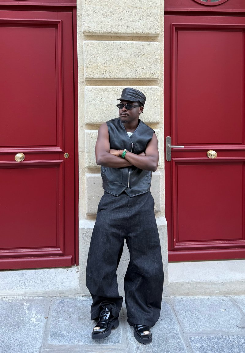 Gilet en cuir noir et pantalon en denim foncé à jambes larges, avec une casquette noire et des lunettes de soleil rondes. Se tenant contre un mur en pierre texturé et des portes rouges.