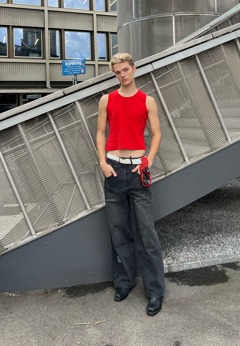 Giovane con top rosso senza maniche e pantaloni neri larghi sta in piedi con le mani nelle tasche vicino a una facciata moderna di un edificio in metallo e vetro.