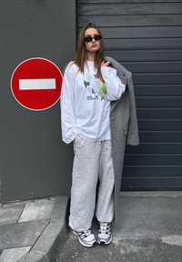 Model wearing a white long-sleeve t-shirt with a graphic design, grey wide-leg sweatpants, and grey oversized coat; black sunglasses and white sneakers.