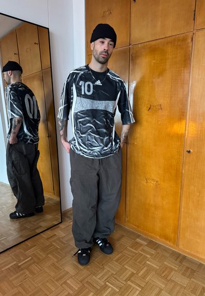 Camiseta Adidas negra y gris con el número "10", pantalones cargo grises sueltos y zapatillas negras con rayas blancas. Fondo de madera y reflejo en el espejo.