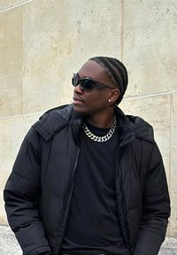 Črna puhovka z kapuco, črna majica, srebrna verižica in temna sončna očala. Oseba stoji ob svetlem, teksturiranem zidu.