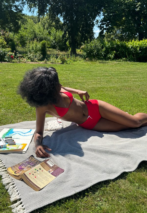 Kvinde i rød bikini ligger på et tæppe i en solrig have med kunstmaterialer og en åben bog i nærheden.