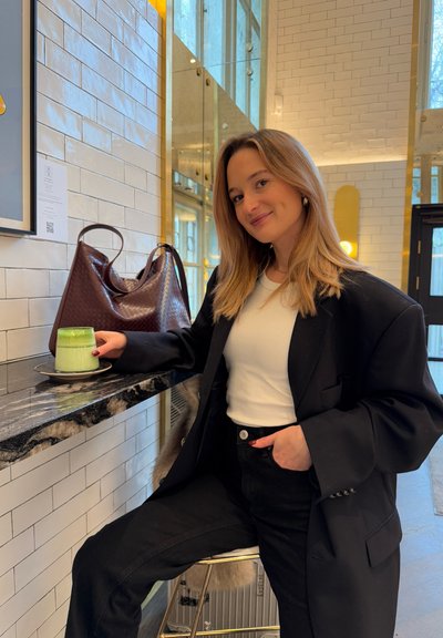 Blazer negro de gran tamaño sobre una camisa blanca, sosteniendo un postre verde en un vaso, sobre una encimera de mármol negro con una bolsa de patrón marrón cerca.