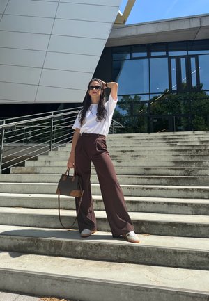 Pantalones de pierna ancha marrones de cintura alta combinados con una camiseta blanca y zapatillas blancas. Un bolso marrón con estampado completa el look.