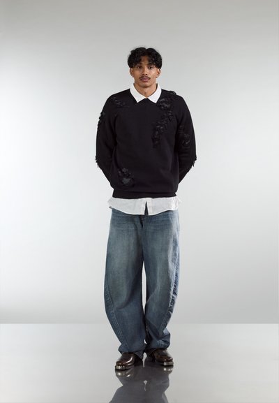 Suéter negro con textura desgastada, camisa de cuello gris claro, jeans azules holgados y zapatos oscuros. Diseño minimalista, sin patrones ni logotipos.