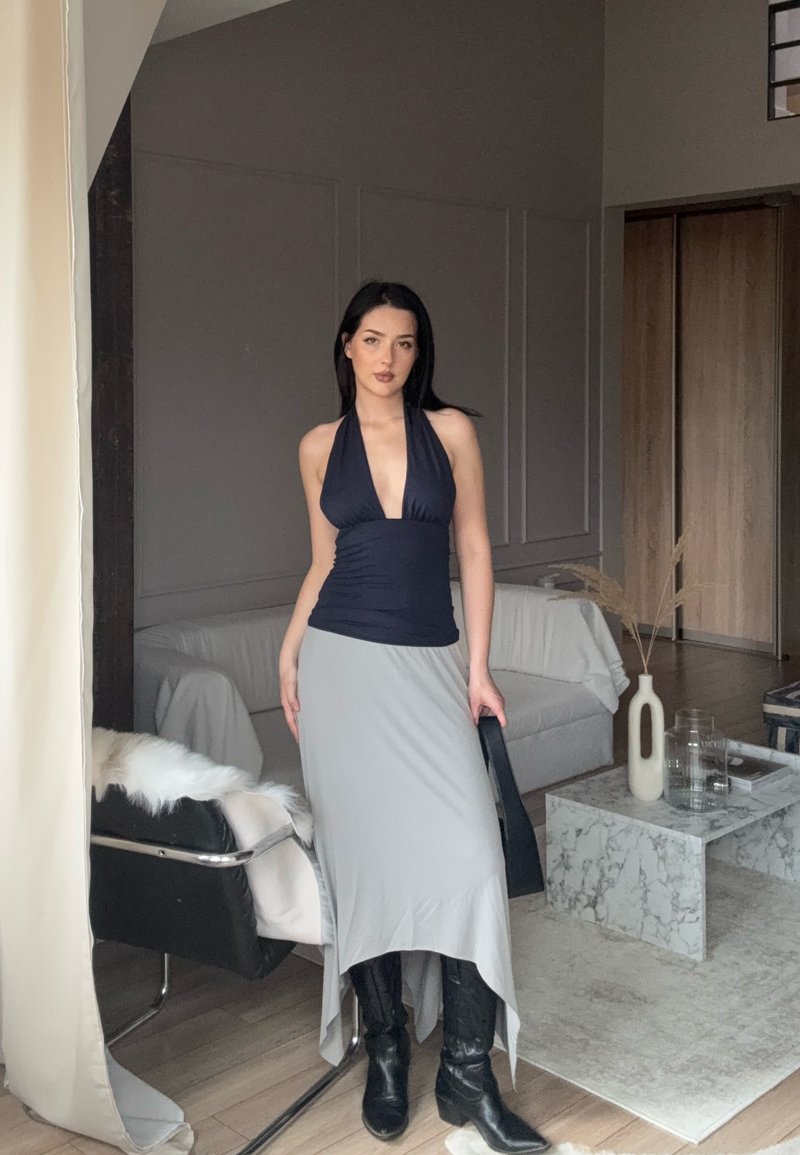 Donna con top nero a collana, gonna asimmetrica grigia e stivali neri che si trova in un soggiorno moderno con tavolino in marmo e divano.