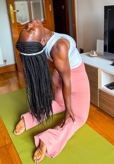 Persona con una camiseta sin mangas gris y pantalones de yoga acampanados rosas realizando una postura de arco sobre una esterilla verde. Pelo largo trenzado, descalza con los dedos de los pies visibles.