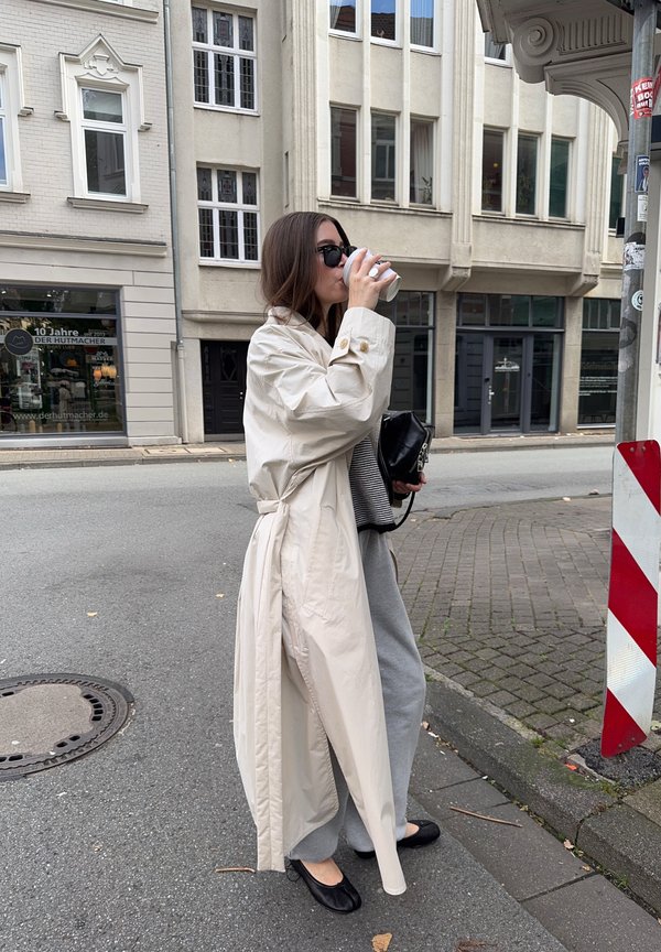 Gabardina beige con cuello ancho y botones, pantalones grises, bolso negro y zapatos planos. Bebiendo de un vaso de papel en un entorno urbano.