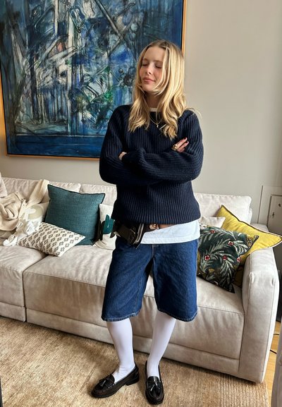 Suéter de punto azul marino, camisa blanca, pantalones cortos de mezclilla, medias blancas, mocasines negros, accesorios que incluyen un cinturón con una funda, en un interior acogedor.