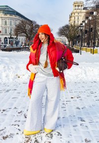 Γυναίκα με κόκκινο μπουφάν, πορτοκαλί καπέλο και κασκόλ στέκεται σε χιονισμένο δρόμο της πόλης με ιστορικά κτήρια στο φόντο.