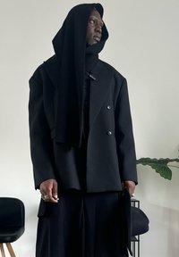 Casaco preto cruzado com ombros oversized, complementado por um cachecol preto texturizado. Modelo segura um chapéu preto, em frente a um fundo claro.