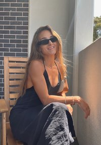 Jeune femme en robe noire et lunettes de soleil assise sur une chaise en bois sur un balcon, souriante avec la lumière du soleil sur le visage et le bras.