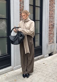 Beiger Strickpullover mit einem Schal, braune weite Hosen und spitze schwarze Absätze, hält eine gewebte schwarze Handtasche. Urbane Umgebung mit Ziegelmauer.