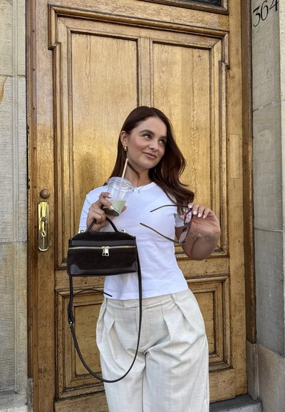 Bolso de ante negro con doble cremallera, combinado con una camiseta blanca y pantalones claros con pliegues. Superficies lisas y hardware minimalista.