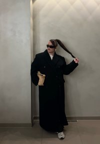 Abrigo largo negro de doble botonadura, camiseta blanca, gafas de sol negras, coleta estilizada, sosteniendo un bolso marrón claro, de pie frente a una pared gris claro.