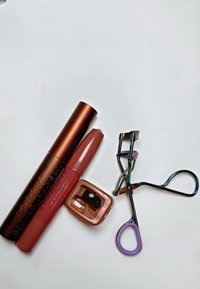 Due tubetti cosmetici: un mascara bronzo scuro e un rossetto rosa opaco, insieme a un piegaciglia metallic rainbow con accenti viola.