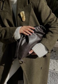 Καφέ δερμάτινη τσάντα με κορδόνι που κρατιέται στα χέρια ενός ατόμου, με ένα υφή πράσινο παλτό και ένα λευκό πουλόβερ ορατά στο φόντο.