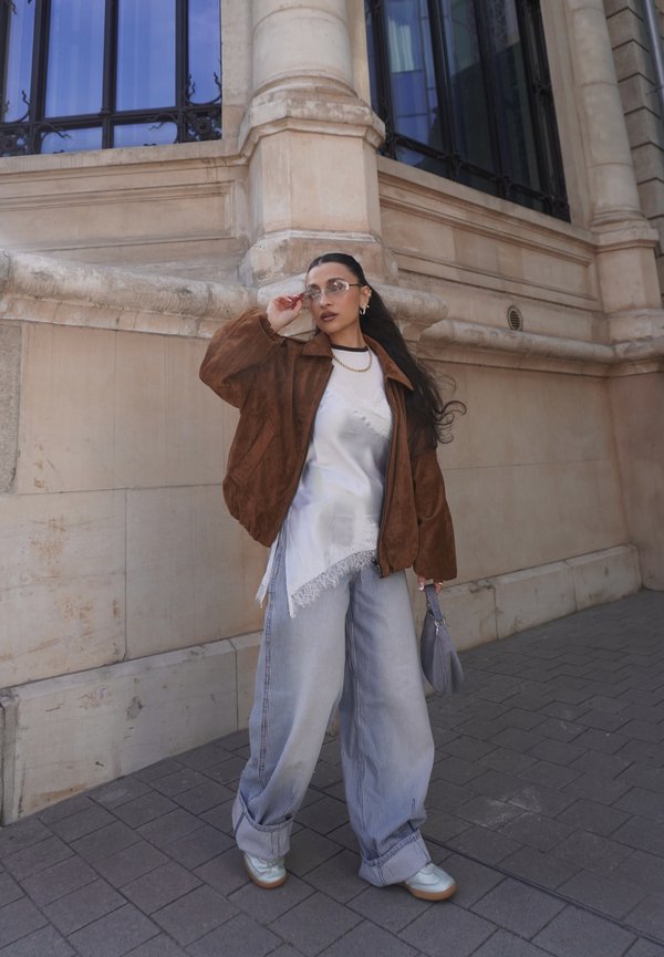 Femme portant une veste marron, un haut blanc asymétrique, un pantalon large à rayures, des chaussures blanches et des lunettes, debout près d'un bâtiment en pierre beige.