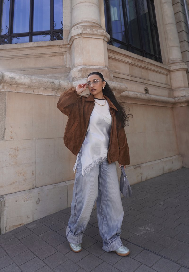 Vrouw met een bruin jasje, witte asymmetrische top, brede gestreepte broek, witte schoenen en een bril, staande naast een beige stenen gebouw.