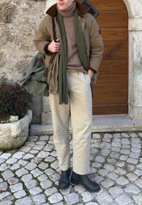 Homme portant un pantalon beige, des bottes noires, un col roulé marron, une écharpe olive et une veste, portant un sac vert debout sur un pavement en pavés.