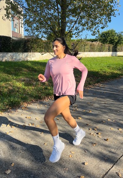 Camisa de running rosa de manga larga, pantalones cortos negros, zapatos y calcetines deportivos blancos. Persona corriendo por un camino con hierba y árboles al fondo.