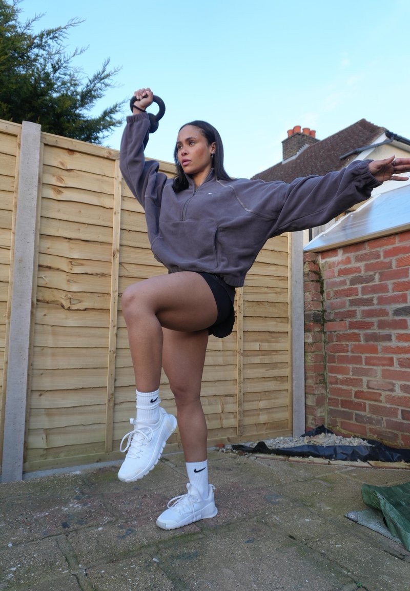 Γυναίκα με αθλητικά ρούχα ισορροπεί σε ένα πόδι σε εξωτερικό χώρο, κρατώντας μια αλτήρα ψηλά με το ένα χέρι εκτεταμένο για σταθερότητα.