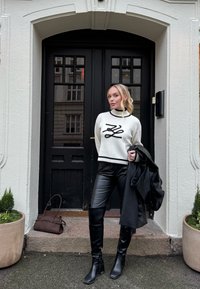 Femme en pull blanc, pantalon en cuir noir et bottes, debout devant une porte double noire, tenant une veste noire, avec un sac brun posé au sol.