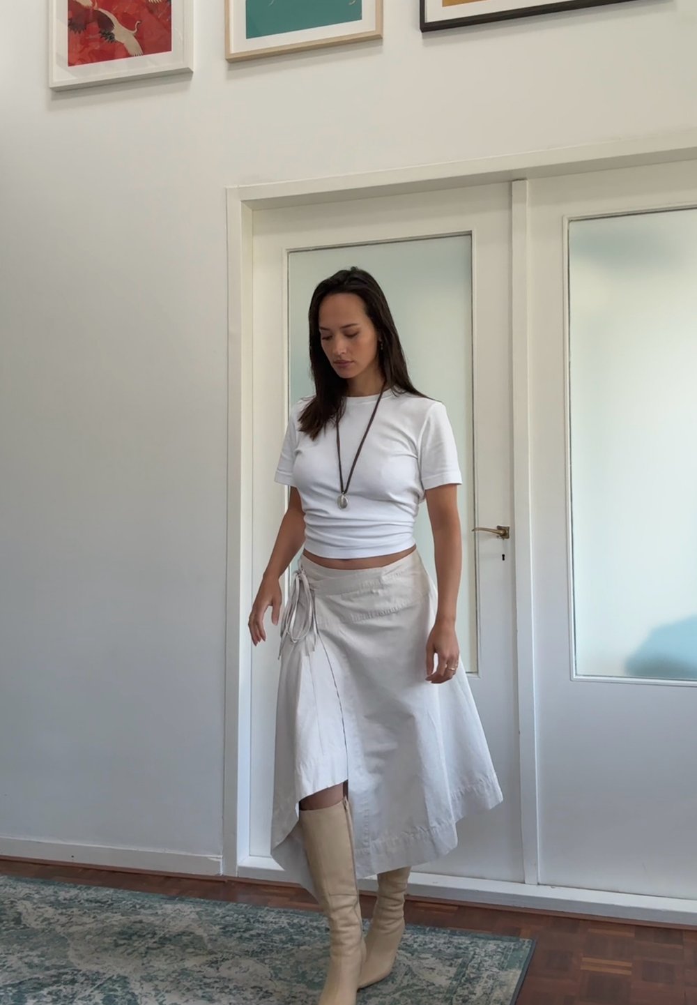 Witte cropped t-shirt, beige omslagrok met strikdetail en kniehoog beige laarzen. Haar is los; op de achtergrond zijn glazen deuren en kunst te zien.