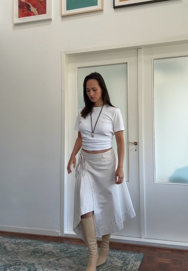 Witte cropped t-shirt, beige omslagrok met strikdetail en kniehoog beige laarzen. Haar is los; op de achtergrond zijn glazen deuren en kunst te zien.