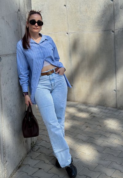 Camisa de botones a rayas azul claro, jeans de mezclilla claros de cintura alta, zapatos negros, cinturón marrón con logo y un bolso oscuro sobre una superficie pavimentada.