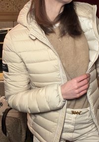 Doudoune beige avec capuche et fermeture éclair, présentant un design matelassé, des poignets côtelés et un écusson logo sur la manche. Portée par-dessus un haut beige clair.