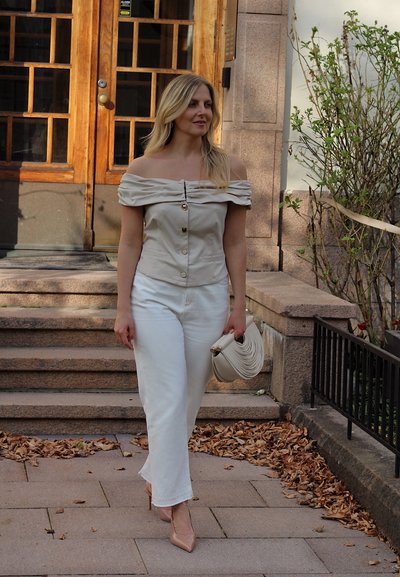 Top beige de hombros descubiertos con botones, combinado con pantalones anchos blancos. Sosteniendo un bolso crema. Escaleras y vegetación de fondo.