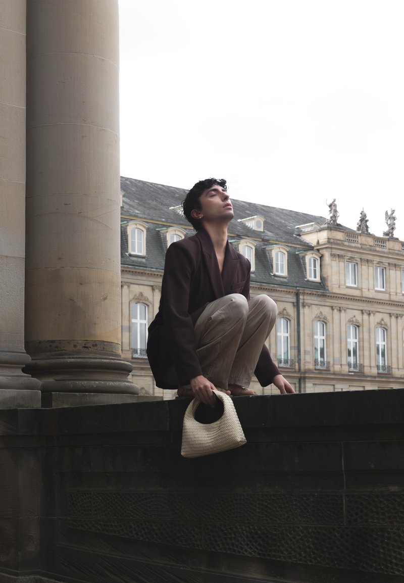 A person squatting on a stone ledge next to a large column, holding a woven bag, with their eyes closed and a historic building in the background under an overcast sky.