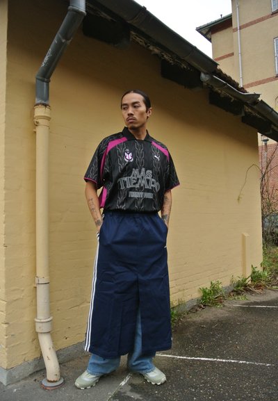 Camiseta de fútbol negra con acentos y rayas rosas, combinada con pantalones cortos amplios azul marino con rayas laterales blancas, llevados con zapatillas verdes.