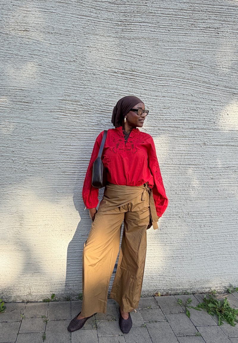 Femme portant un chemisier rouge, un pantalon marron à jambes larges, un foulard noir sur la tête et des lunettes, se tenant contre un mur texturé gris clair, regardant vers la droite.