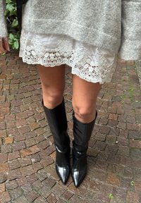 Čierne čižmy po kolená s lesklým povrchom a špicatou špičkou, nosené s bielou čipkovanou sukňou, stojace na štruktúrovanej dlažbe z kociek.