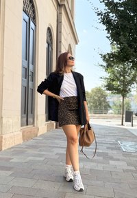 Μαύρο σακάκι πάνω από λευκό T-shirt, φούστα με λεοπάρ σχέδιο και ανακλαστικά αθλητικά παπούτσια. Καφέ τσάντα χειρός, στέκεται σε πέτρινη πλακόστρωτη επιφάνεια.