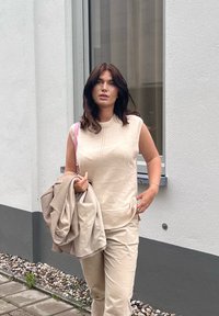 Sieviete ar tumšiem matiem, tērpusies bēša krāsas bezpiedurknu džemperī un biksēs, nēsā bēša krāsas jaku, ejot pa ietvi gar baltas ēkas sienu.