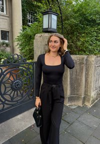 Una persona con una maglietta nera a maniche lunghe e pantaloni neri a vita alta tiene in mano una piccola borsa nera, in piedi vicino a un muro di pietra e alla vegetazione.
