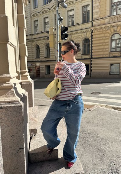 Mujer con gafas de sol, camisa a rayas y vaqueros anchos bebe una bebida mientras está de pie en los escalones de la acera de la ciudad cerca de un semáforo.