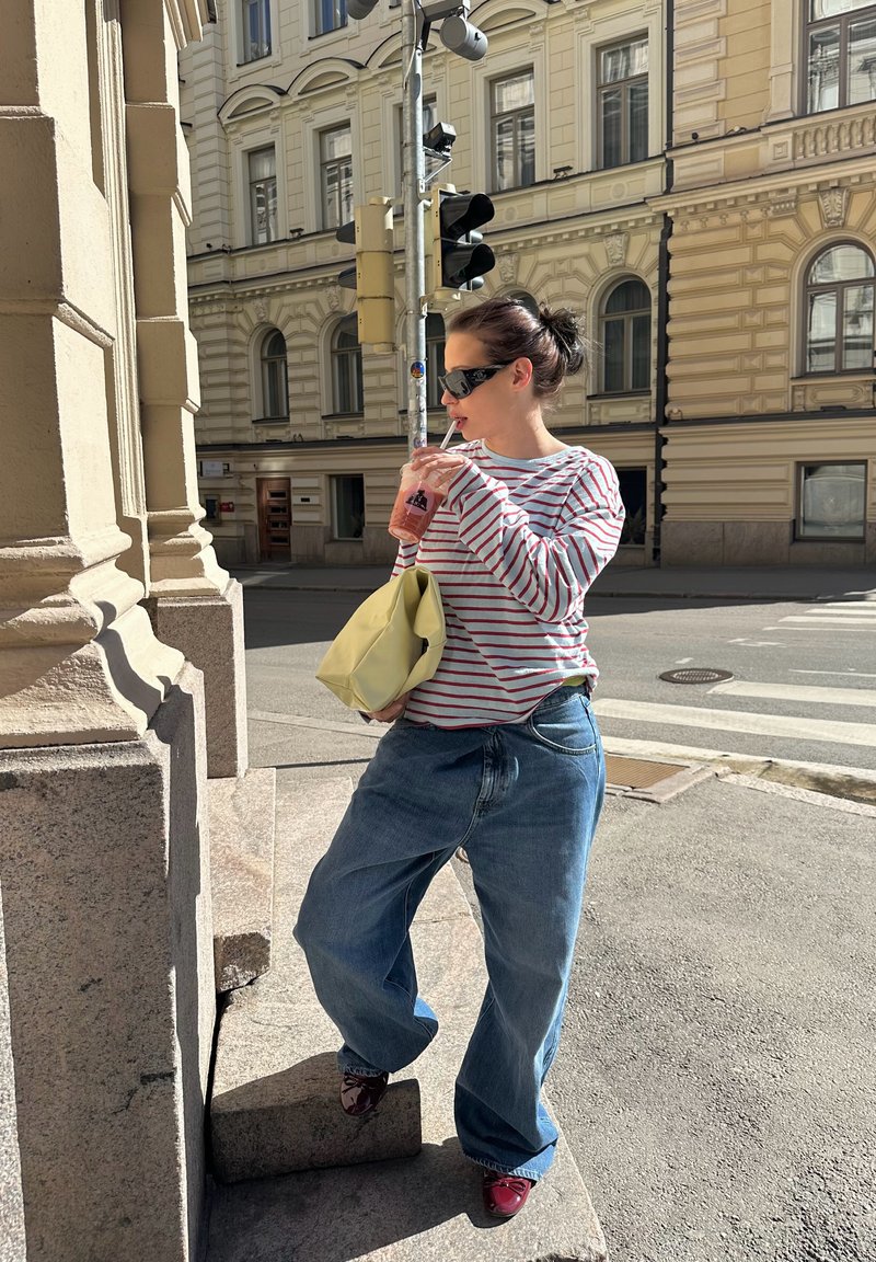 Femme portant des lunettes de soleil, une chemise rayée et un jean large, sirote une boisson en se tenant sur les marches du trottoir de la ville près du feu de circulation.