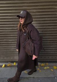 Barna gyapjú kabát nagy gombokkal, sötétbarna velúr táska, magas csizma, sötét sapka és napszemüveg; meleg sál a vállakat takarva.