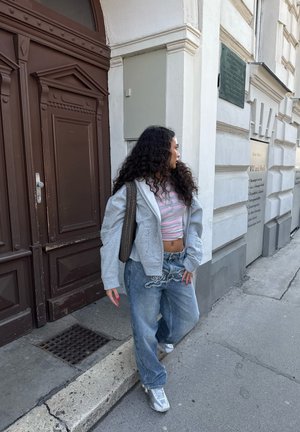 Mujer con cabello largo y rizado que lleva un crop top a rayas, sudadera gris con capucha y vaqueros anchos, de pie en la acera cerca de una puerta marrón y un edificio blanco.