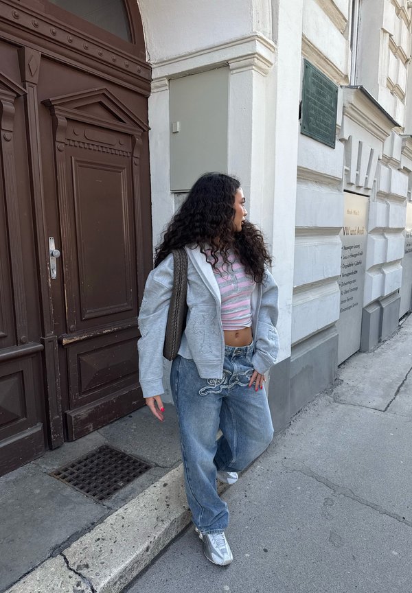Žena s dlhými kučeravými vlasmi v pruhovanom crop tope, sivej kapucni a voľných džínsach stojí na chodníku blízko hnedých dverí a bielej budovy.