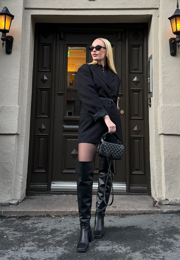 Vestido blazer negro con cinturón, medias negras transparentes, botas negras por encima de la rodilla y bolso negro acolchado. Texturas mate y diseño elegante.