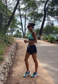 Αθλητική ενδυμασία: τιρκουάζ αθλητικό σουτιέν, μαύρη κοντή παντελόνι με λευκά σχέδια και πολυχρωμα παπουτσάκια τρεξίματος. Φυσικό φόντο με δέντρα.