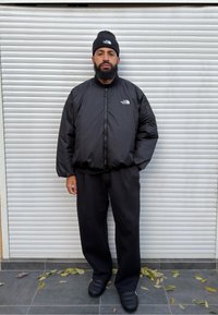 Sort dunjakke med lynlås og logo på brystet, sort beanie, løse sorte bukser og mønstrede sorte sko. Overflade med blade.