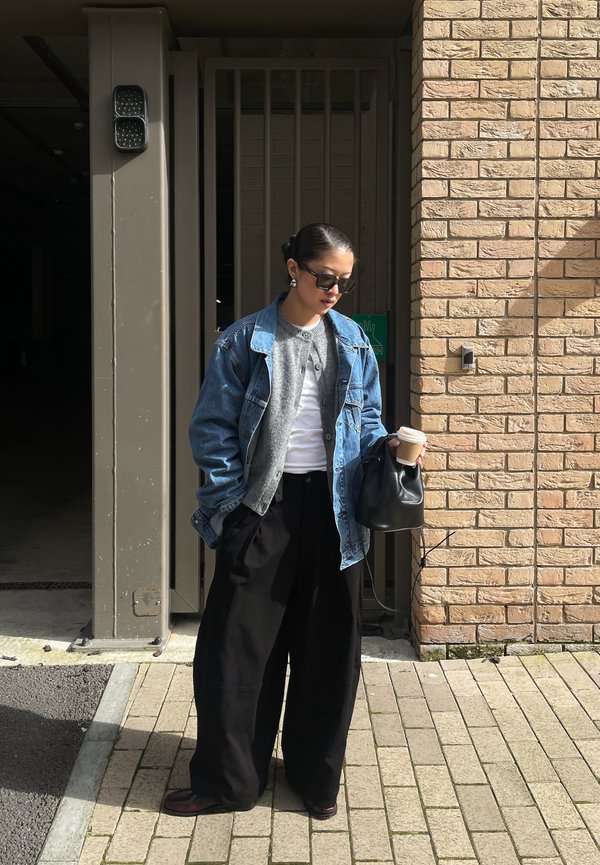 Femme portant des lunettes de soleil, une veste en denim, un cardigan gris et un pantalon large noir, tenant une tasse de café et un sac noir près d'un mur de briques.